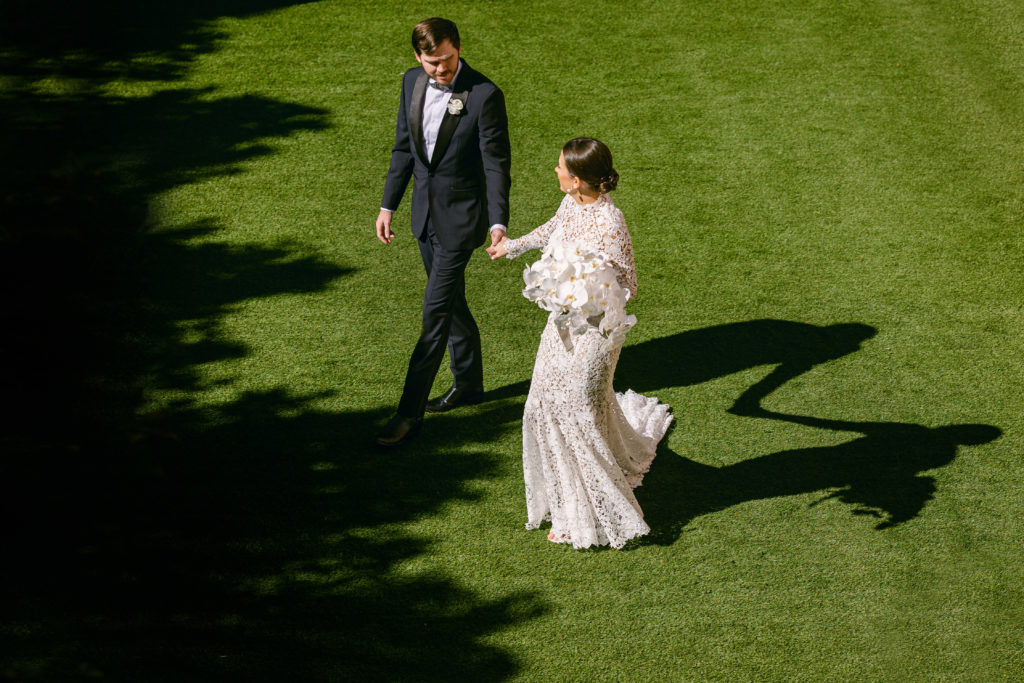 Bride in her patterned lace wedding dress and groom in a black tux pose for a picture on the Event lawn at the Rosewood Mansion on Turtle creek during their wedding photographed by Carter Rose