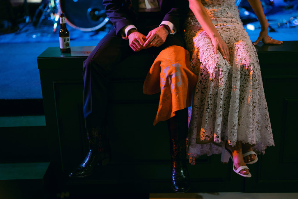 Wedding couple sit together after the reception. The groom is wearing a black tux and cowboy boots and the bride is wearing a delicate patterned lace dress.